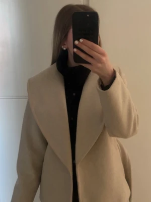Beige kappa från Vera Moda  - Stilren beige kappa med bred krage och knytskärp i midjan. Kappan har en rak passform och är tillverkad i ett mjukt, ull-liknande material. Perfekt för att hålla värmen under kyliga dagar och ger en snygg siluett. Kappan kommer inte till användning 