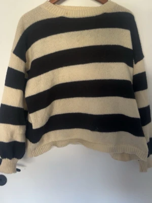 Randig stickad tröja by clara Paris - Stickad tröja från by clara Paris med breda svarta och beige ränder. Tröjan har rund halsringning, långa ärmar och ribbade muddar. Perfekt för dig som gillar en avslappnad och trendig look.