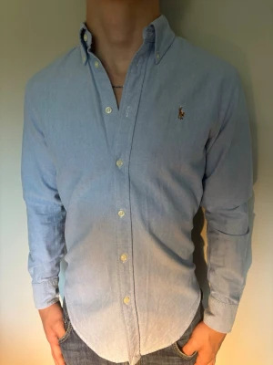 Ljusblå skjorta från Ralph Lauren - Klassisk ljusblå skjorta från Ralph Lauren med button-down krage och broderad logga på bröstet. Skjortan har långa ärmar, knappar framtill och är tillverkad i mjuk bomull. Perfekt för en stilren och avslappnad look. Riktigt schysst till sommaren!
