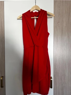 Röd omlottklänning med v-ringning - Snygg röd kortklänning med omlott och djup v-ringning. Klänningen är ärmlös och har en elegant detalj i ryggen med öppning. Perfekt för dig som vill sticka ut med en klassisk och stilren look.