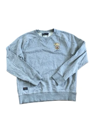 Grå sweatshirt från Morris - Grå sweatshirt från Morris med broderad logga och klubbmärke på bröstet. Tröjan har rund halsringning, långa ärmar och ribbade muddar. Perfekt för en sportig och avslappnad stil.