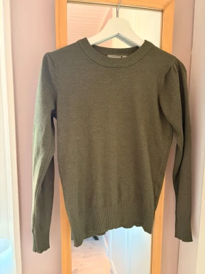 Olivgrön stickad tröja med puffärm - Snygg olivgrön stickad tröja med rund halsringning och diskreta puffärmar. Tröjan har ribbstickade muddar vid ärmslut och nederkant, vilket ger en stilren och modern look. Perfekt att matcha med jeans eller kjol.