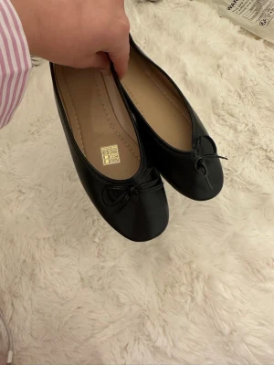 Svarta ballerinaskor med rosett - Stilrena svarta ballerinaskor från H&M med klassisk rund tå och söt rosettdetalj framtill. Skorna har en slät ovandel i syntetmaterial och beige innersula. Perfekta för dig som gillar enkel och tidlös stil.