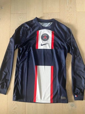 PSG långärmad tröja Nike - Snygg långärmad tröja från Paris Saint-Germain i mörkblått med vita och röda detaljer. Tröjan har PSG-logga och Nike Swoosh på bröstet samt Dri-FIT-material som andas. Perfekt för fotbollsträning eller att bära som supporter. Den är helt ny och oanvänd.