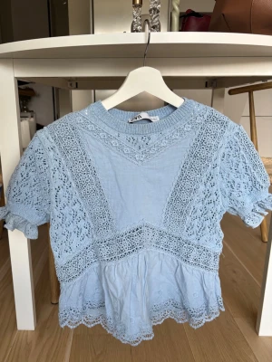 Ljusblå spetsblus från Zara - Superfin ljusblå blus från Zara med vackra spetsdetaljer och korta ärmar. Blusen har rund halsringning och volangkant nertill, perfekt för en söt och trendig look. Luftigt material som passar bra till jeans eller kjol.