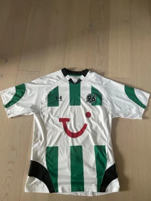 Hannover 96 matchtröja Under Armour - Vit och grön fotbollströja från Hannover 96 med svarta detaljer och klubbens logga på bröstet. Det är Hannoverns borta tröja säsongen 2009-2010. Klassisk passform med korta ärmar och rund hals. Det är en  gul fläck ganska centralt på framsidan men kan nog tas bort om man försöker, ingen garanti. Den är köpt på en pop up butik som bara säljer retro fotbollströjor.