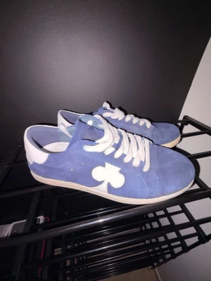 The Gallerist - Snygga blå sneakers i mocka med vit klöverdetalj på sidan och vita skosnören. Skorna har en låg siluett och vit sula, samt vit hälkappa. Perfekta för dig som vill sticka ut med en unik detalj på dina skor. Nästan helt nya, använder ej mycket då jag köpte nya skor, priset kan diskuteras!