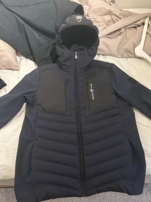 Västjacka - Sail Racing - Snygg mörkblå västjacka från Sail Racing med huva och hel dragkedja. Jackan har två bröstfickor med dragkedja och logga på bröstet.