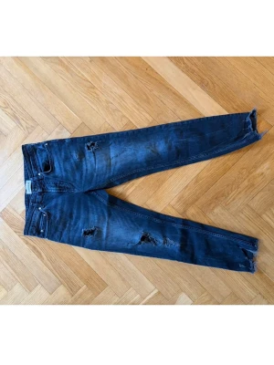 Mörkblå slitna jeans  - Snygga mörkblå jeans med slitningar och hål på benen. Jeansen har klassisk femficksdesign och råa benslut för en trendig look. Perfekta för dig som gillar en avslappnad och edgy stil.