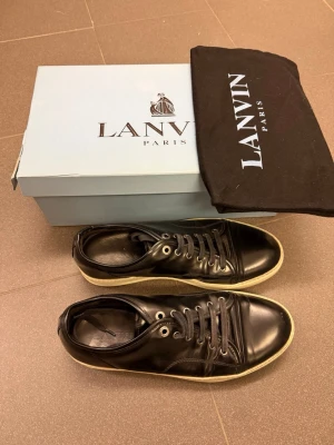 Lanvins - Stilrena svarta sneakers från Lanvin i blankt läder med vita sulor och svarta skosnören. Klassisk låg modell med rund tå och diskreta sömmar. Kommer med originalkartong och dustbag. Perfekta för dig som gillar lyxiga och tidlösa sneakers.