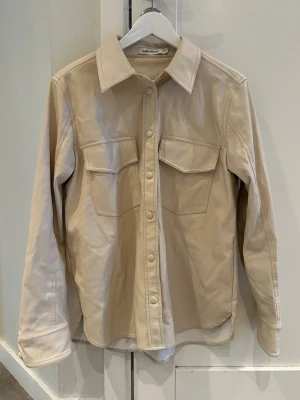 Beige overshirt från Carin Wester - Stilren beige overshirt i skinnliknande tyg från Carin Wester. Overshirten har klassisk krage och är tillverkad i ett slätt material som ger en clean look. Perfekt att bära öppen eller stängd för en trendig outfit.