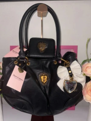 Juicy Couture svart handväska - Svart handväska från Juicy Couture i mjukt konstläder med guldfärgade detaljer och emblem framtill. Väskan har dubbla handtag, dekorativ vit rosett och hjärtformad nyckelring. Perfekt för dig som gillar en trendig och klassisk look.