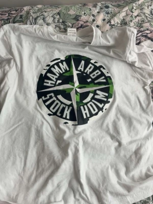Hammarby t-shirt  - Vit t-shirt med stort Hammarby Stockholm-tryck i svart och grönt på bröstet. Klassisk rund hals och korta ärmar. Perfekt för dig som vill visa ditt stöd för Hammarby på ett stilrent sätt.