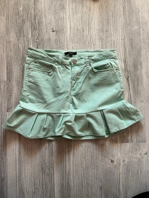 Mintgrön volangkjol från Lindex  - Supersöt mintgrön kortkjol från lindex med volangkant nedtill. Kjolen har klassiska jeansfickor fram och bak, bälteshällor och stängs med knapp och dragkedja. Perfekt för dig som vill ha en fräsch och lekfull look. (Kjolen är grön/coral blå)