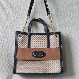 Beige och brun handväska med kedja - Snygg handväska i beige och brunt med svart kant och guldfärgade kedjor. Väskan har ett grafiskt mönster, dubbla handtag och en axelrem. Framtill sitter en dekorativ detalj i svart och guld. Perfekt för dig som gillar trendiga accessoarer.