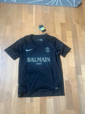 Svart PSG x Balmain tröja Nike - Snygg svart t-shirt från Nike i samarbete med PSG och Balmain. Tröjan har rund hals, korta ärmar och tryck med Balmain Paris samt PSG-logga på bröstet. Tillverkad i Dri-FIT material som andas, perfekt för träning eller sport.