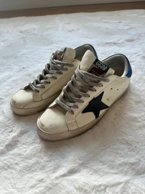 Golden Goose vita sneakers med blå detalj - Golden Goose sneakers i vitt läder med svart stjärna på sidan och blå hälkappa. Gråspräckliga skosnören och GGDB-logga på plösen. Klassisk låg modell med slitna detaljer för en cool, avslappnad look.