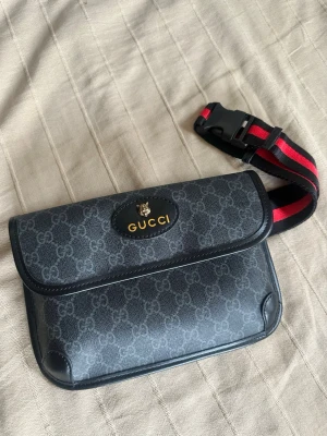 Svart axelväska  - (OBS VET EJ OM DEN ÄR ÄKTA!!!!) Snygg svart axelväska med klassiskt GG-mönster och guldfärgad logga framtill. Väskan har en justerbar axelrem i svart och rött med spänne. Tillverkad i canvas med läderdetaljer och stilren, rektangulär form.