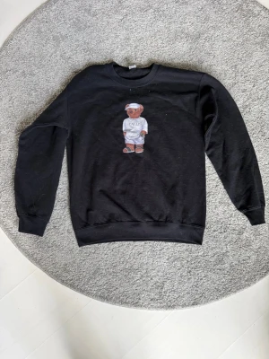 Svart sweatshirt med björntryck  - Cool svart sweatshirt från Gildan med ett tryck av en björn i mitten som har på sig en vit tröja och keps. Tröjan är i modellen Heavy Blend och har rund hals samt långa ärmar. Perfekt för dig som gillar streetwear vibes.
