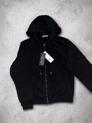 Svart hoodie från C.P. Company - Svart hoodie med dragkedja från C.P. Company. Klassisk modell med huva, ribbade muddar och fickor framtill. Ärmen har den ikoniska linsen med C.P. Company-loggan. Perfekt för en stilren och avslappnad look.