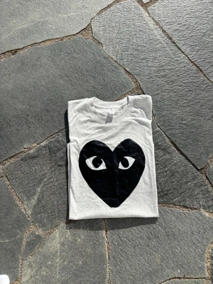 COMME des GARÇONS - Snygg grå t-shirt med ett stort svart hjärta med ögon på bröstet. T-shirten har rund hals och korta ärmar, perfekt för en cool och avslappnad stil. Passar dig som gillar grafiska tryck och vill sticka ut lite extra.