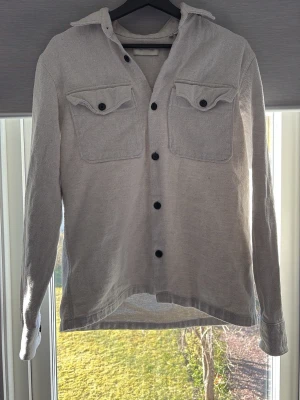 Ljusgrå overshirt med fickor - Snygg ljusgrå overshirt med svarta knappar och två bröstfickor med lock. Overshirten har klassisk krage, långa ärmar och är tillverkad i ett mjukt, strukturerat tyg. Perfekt att bära öppen eller stängd för en avslappnad stil.