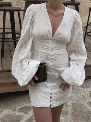 Vit broderad klänning från Adore - Superfin vit klänning från Adore med broderade blommor, djup v-ringning och långa puffärmar. Klänningen har knappar framtill och är figurnära med kort längd. Perfekt för dig som gillar romantisk och trendig stil.