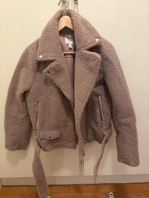 Rosa teddyjacka med bälte - Mysig teddyjacka i ljusrosa med bred krage, dragkedjor och bälte i midjan. Jackan har två fickor med dragkedja och en cool bikerinspirerad design. Perfekt för kalla dagar när du vill vara både varm och snygg.