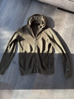 Olivgrön hoodie med dragkedja - Säljer en olivgrön hoodie med hel dragkedja och två fickor framtill. Huvan är fodrad med ett glansigt material som ger en schysst kontrast till den stickade strukturen på resten av tröjan. Perfekt för dig som gillar en avslappnad och stilren look.