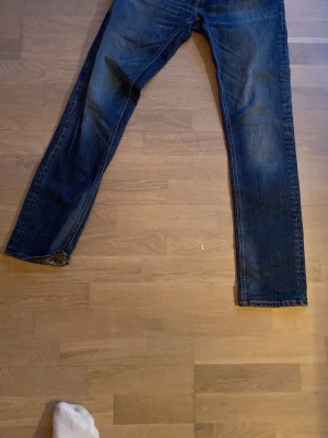 Mörkblå Nudie jeans  - Säljer ett par mörkblå jeans med raka ben och klassisk femficksdesign. Jeansen har lätt slitna detaljer och är tillverkade i denim. Perfekta för en avslappnad och stilren look.