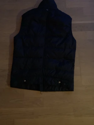 Mörkblå J.Lindeberg Väst  - Snygg svart dunväst med hög krage och dragkedja framtill. Västen har en quiltad design och knappar längst ner bak. Perfekt för lager på lager och håller dig varm under kyliga dagar.