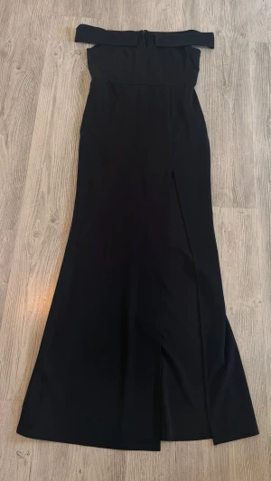 Svart axelbandslös långklänning - Elegant svart långklänning med axelbandslös design och figurnära passform upptill. Klänningen har en lätt utsvängd nederdel och en stilren look, perfekt för mer uppklädda tillfällen.