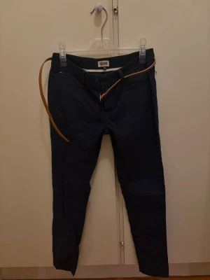 Mörkblå chinos från Tommy Hilfiger - Snygga mörkblå chinos från Tommy Hilfiger med smal passform och klassisk design. Byxorna har två framfickor, två bakfickor och medföljer ett smalt brunt bälte. Perfekta för en stilren look.