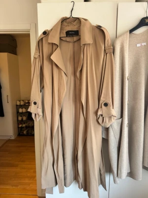 Beige trenchcoat från vero moda - Stilren beige trenchcoat från vero moda med klassisk krage, axelklaffar och knappar vid ärmslut. Lång modell med avslappnad passform och snygga detaljer som ger en tidlös look. Perfekt för lager-på-lager och enkel att matcha med olika outfits.det finns en pytte liten svart prick back mitt rygg. Se sista bilden