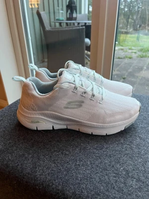 Vita Skechers Arch Fit sneakers - Vita Skechers Arch Fit sneakers. Fräscha vita sneakers från Skechers med Arch Fit-sula för extra komfort. Skorna har en låg profil, mesh-överdel och diskret logga på sidan. Perfekta för dig som gillar stilrena och lätta sneakers med modern känsla.