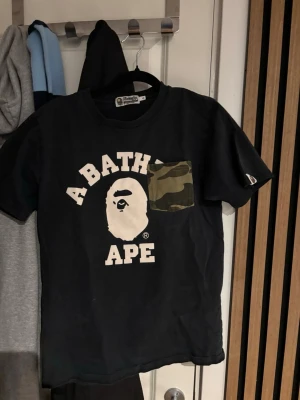 Svart BAPE t-shirt med camoficka - Säljer en svart t-shirt från A Bathing Ape med ikoniskt tryck framtill och en grön camouflagemönstrad bröstficka. Klassisk rund hals och korta ärmar. Perfekt för dig som gillar streetwear och vill sticka ut. Köpt i Bape butiken!
