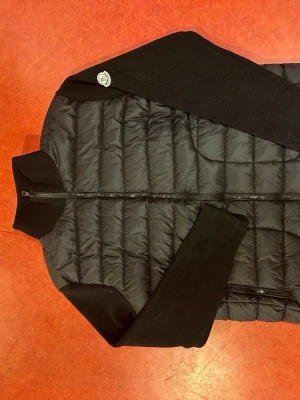 Svart pufferjacka  från Moncler - Svart dunjacka från Moncler med quiltad framsida och stickade svarta ärmar och axelpartier. Jackan har dragkedja framtill och Moncler-logga på axeln. Perfekt för dig som gillar stilren och sportig look.