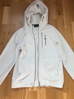 Vit hoodie från Sail Racing - Vit hoodie från Sail Racing med dragkedja framtill och en bröstficka med logga. Hoodien har en stor huva med tryckt detalj och är tillverkad i mjukt material.