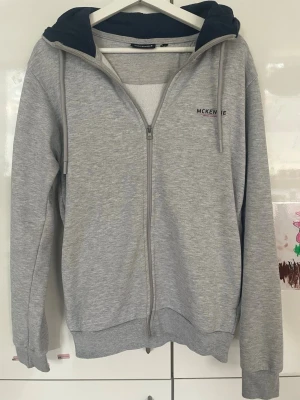 Grå zip-hoodie från McKenzie - Grå hoodie med dragkedja från McKenzie. Mörkblått foder i huvan, ribbade muddar och nederkant. Liten logga på bröstet. Perfekt för lager-på-lager och chill dagar. Klassisk och enkel design som funkar till det mesta.