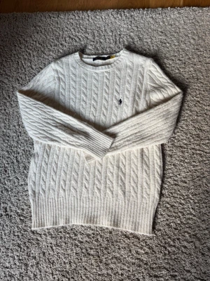 Vit kabelstickad tröja Polo Ralph Lauren - Kabelstickad vit tröja från Polo Ralph Lauren med rund halsringning och broderad marinblå logga på bröstet. Tröjan har långa ärmar och ribbstickade muddar samt nederkant.                                                                            Tröjan är i väldigt fint skick och så men tyvärr så gick jag in i en spik för längesen vilket råka dra upp en tråd på höger arm men det går definitivt att fixa 👍🏽(pris kan diskuteras)