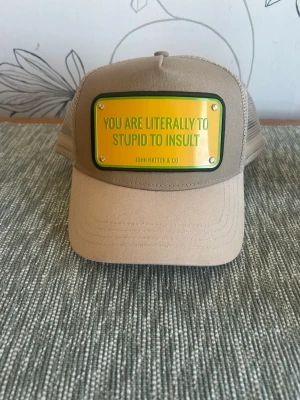 Beige keps John Hatter & Co - Snygg helt oanvänd beige keps från John Hatter & Co med mesh på sidorna och baksidan. Framtill sitter en gul skylt med texten 'YOU ARE LITERALLY TO STUPID TO INSULT'. 380 kr och som sagt helt oanvänd 