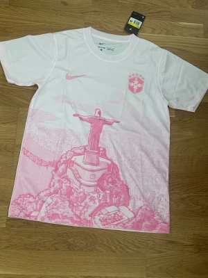 Vit och rosa Nike t-shirt Brasilien - Nike t-shirt i vitt och rosa med tryck av Kristusstatyn i Rio och brasiliansk sköld på bröstet. Klassisk passform med korta ärmar och rund hals. Perfekt för dig som gillar sportig stil och snygga detaljer.