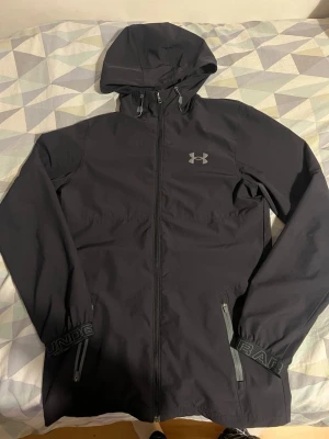 Svart vindjacka från Under Armour - Snygg svart vindjacka från Under Armour med huva och dragkedja framtill. Jackan har två sidofickor med dragkedja, justerbar huva och logga på bröstet. Perfekt för blåsiga dagar och sportiga outfits.