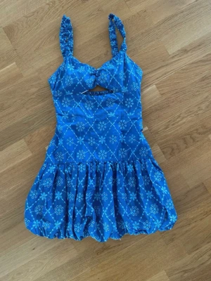  Ny Blå kort klänning med mönster - Söt blå kortklänning med vita blommiga mönster och ballongkjol. Klänningen har breda axelband, liten cut-out framtill och smock i ryggen för extra komfort. Perfekt för varma dagar och somriga tillfällen.