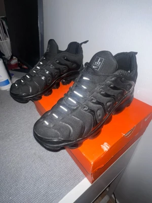 Nike Air VaporMax  - Nike Air VaporMax Plus i helsvart med reflekterande detaljer på snörningen. Ovandel i textil och syntet med vågformade paneler och en unik, synlig VaporMax-sula för extra komfort och stil. Perfekta för dig som gillar en sportig och modern look. Använd ett par gånger. Skorna är rena utanpå och har inga hål i bubblorna. Väldigt bra skick, bara tyget på hälen har öppnats lite men de märks inte 