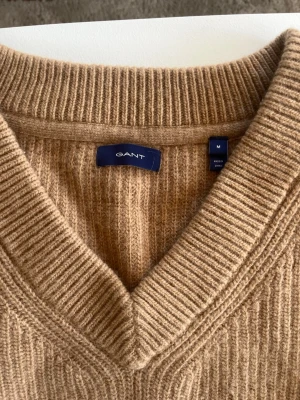 Beige stickad tröja från GANT - Snygg beige stickad tröja från GANT med djup V-ringning och breda ribbade muddar. Tröjan är i mjukt material och har en klassisk, avslappnad passform som passar perfekt till jeans eller kjol.