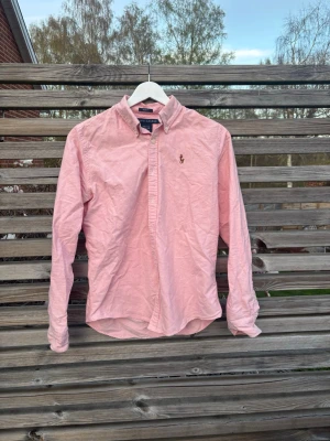 Rosa skjorta från Ralph Lauren - Klassisk rosa skjorta från Ralph Lauren med button-down krage och broderad logga på bröstet. Skjortan har lång ärm, knappar framtill och är tillverkad i bomull. Perfekt för en stilren och avslappnad look. Köpte den för 600kr för några år sen och säljer den nu för den inte passar längre. Hör av er vid frågor!