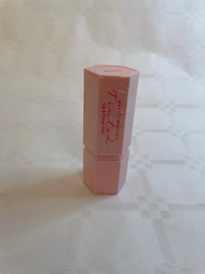 Tinted Lip & Cheek Balm Acai - Säljer ett Tinted Lip & Cheek Balm från Makeup Mekka i färgen Acai. Förpackningen är ljusrosa och har en sexkantig form. Perfekt för dig som vill ha både färg och vård till läpparna. Enkel att ta med i väskan.