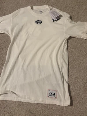 Vit R9 t-shirt från dna NIKE - Snygg vit t-shirt från dna med R9-logga broderad på bröstet och en patch nertill. Klassisk rund hals och korta ärmar. Tillverkad i mjukt bomullsmaterial som känns skönt mot huden. Perfekt för en clean och sportig look.