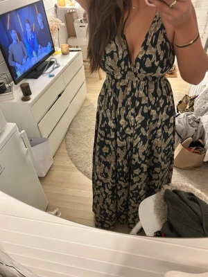 långklänning - Snygg långklänning med svart bas och beige abstrakt mönster. Djup v-ringning och smala axelband ger en trendig look. Klänningen har markerad midja och ett luftigt fall, perfekt för varma dagar. Materialet känns lätt och bekvämt mot huden. Perfekt nu när balen är i säsong